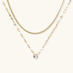 Layered gold necklace with herringbone and link chains, small bezel-set crystal pendant on cream background