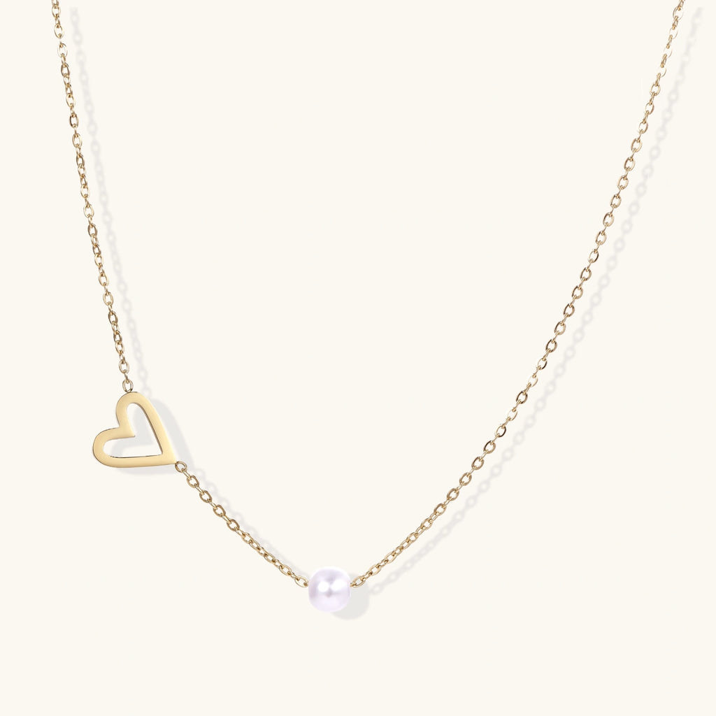 Delicate gold necklace with open heart charm and white pearl bead on fine chain against cream background