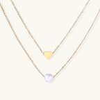 Two delicate gold-plated chains with heart charm and white pearl bead on cream background, minimal layered design.