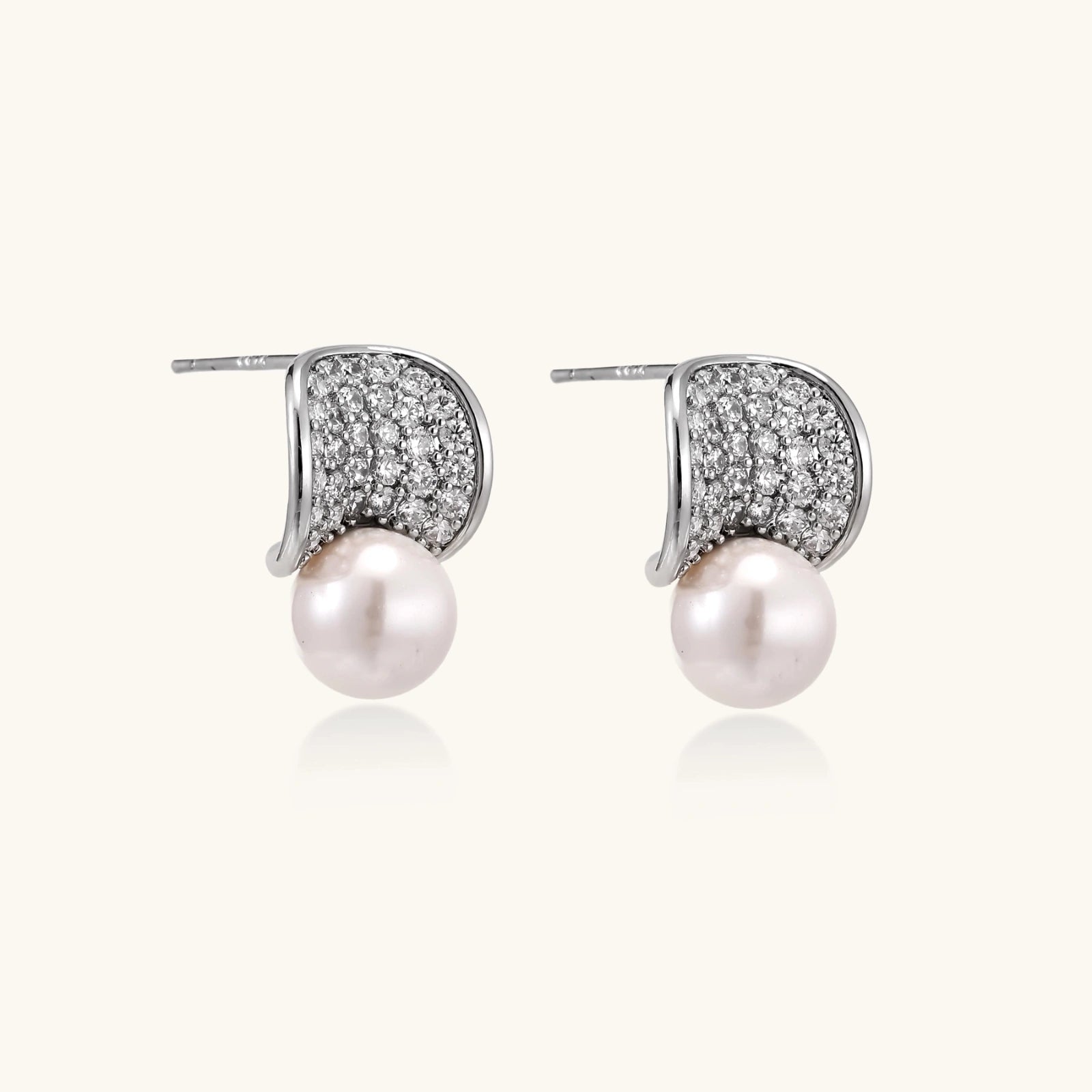Curved pavé diamond studs in white gold-plated brass cradle lustrous freshwater pearls against cream background