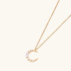 Gold crescent moon pendant with graduated white pearls on delicate chain against cream background
