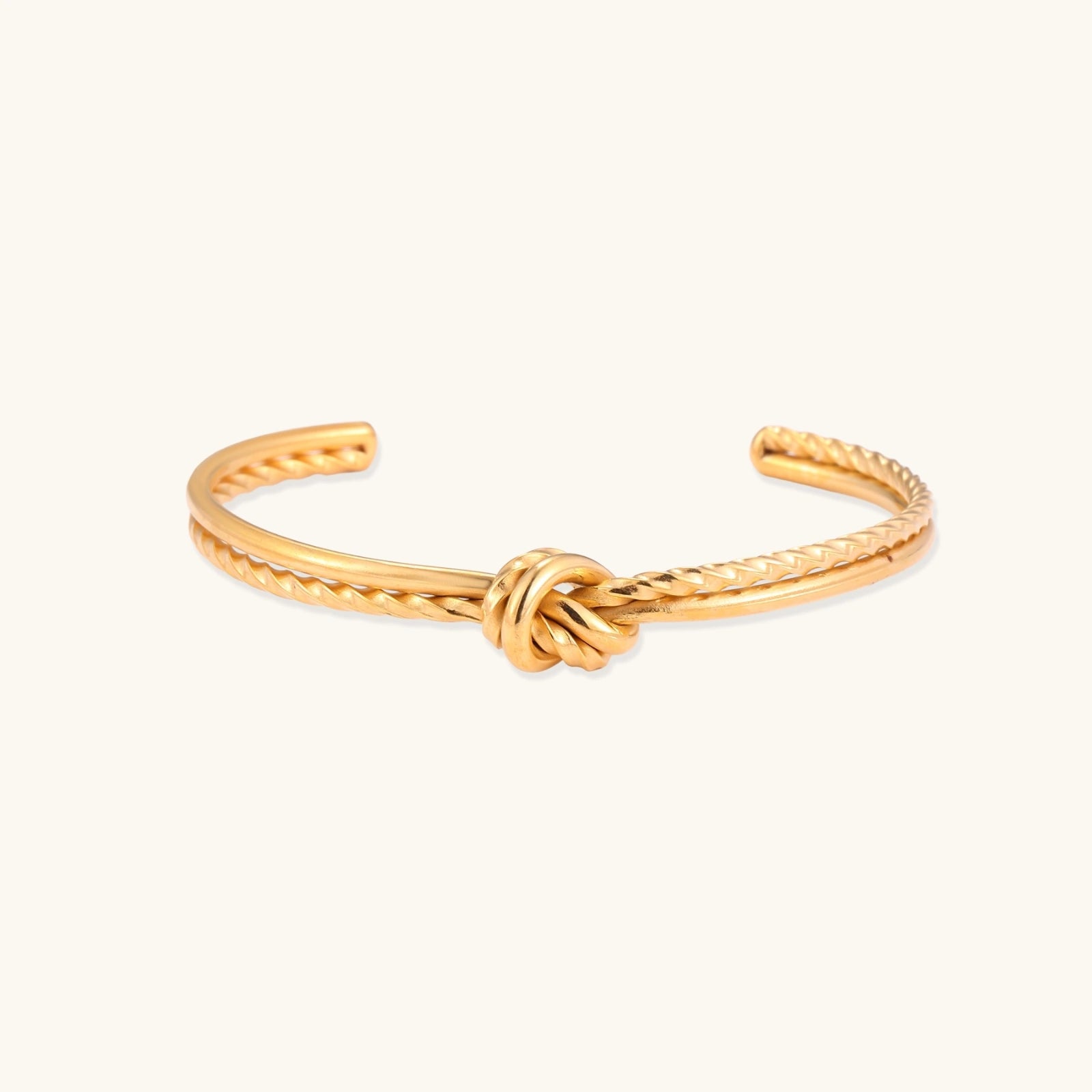 Gold cuff bracelet with twisted rope texture and decorative knot centerpiece on cream background.