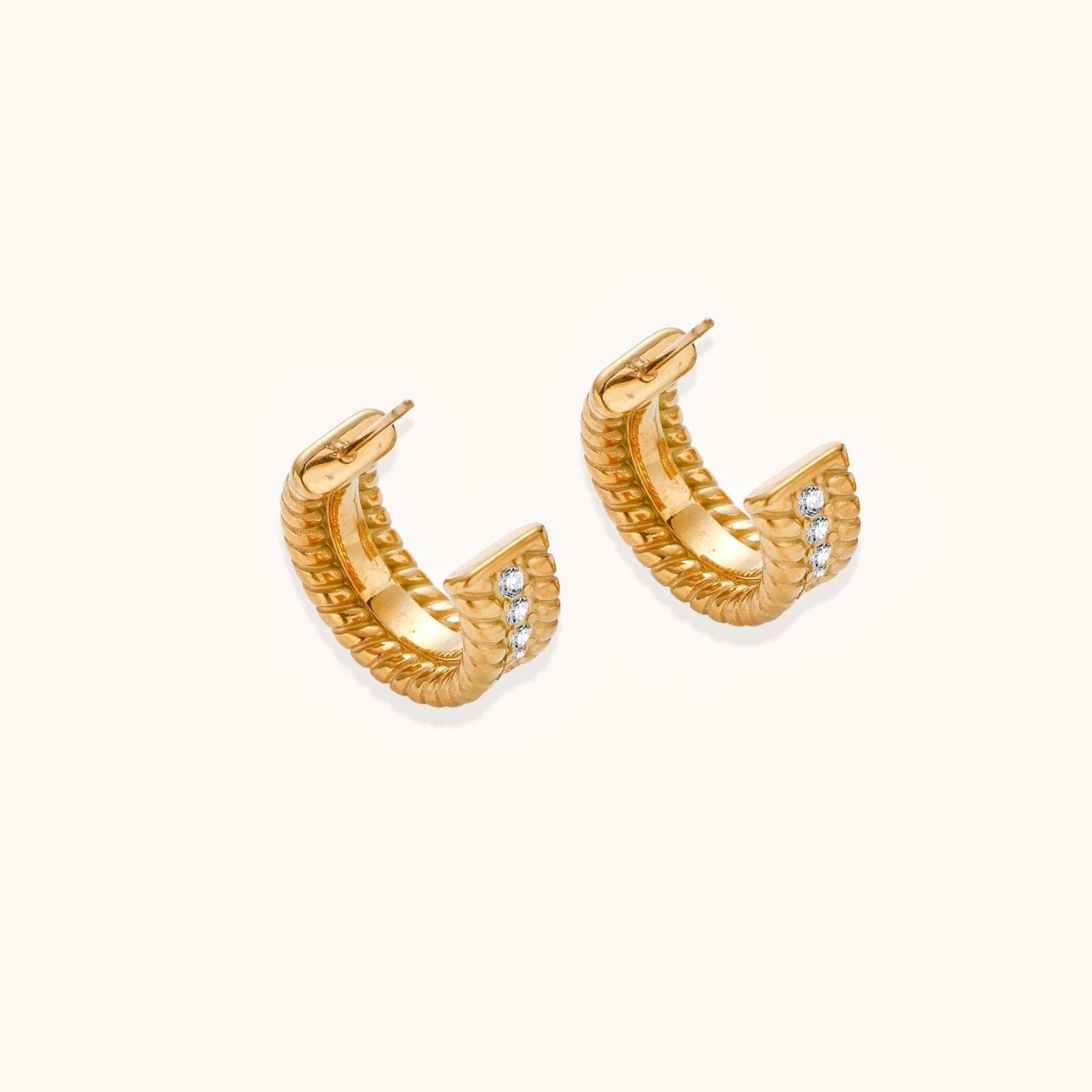 Textured gold huggie hoop earrings with raised ribbed rows creating dimensional sculptural design on cream background